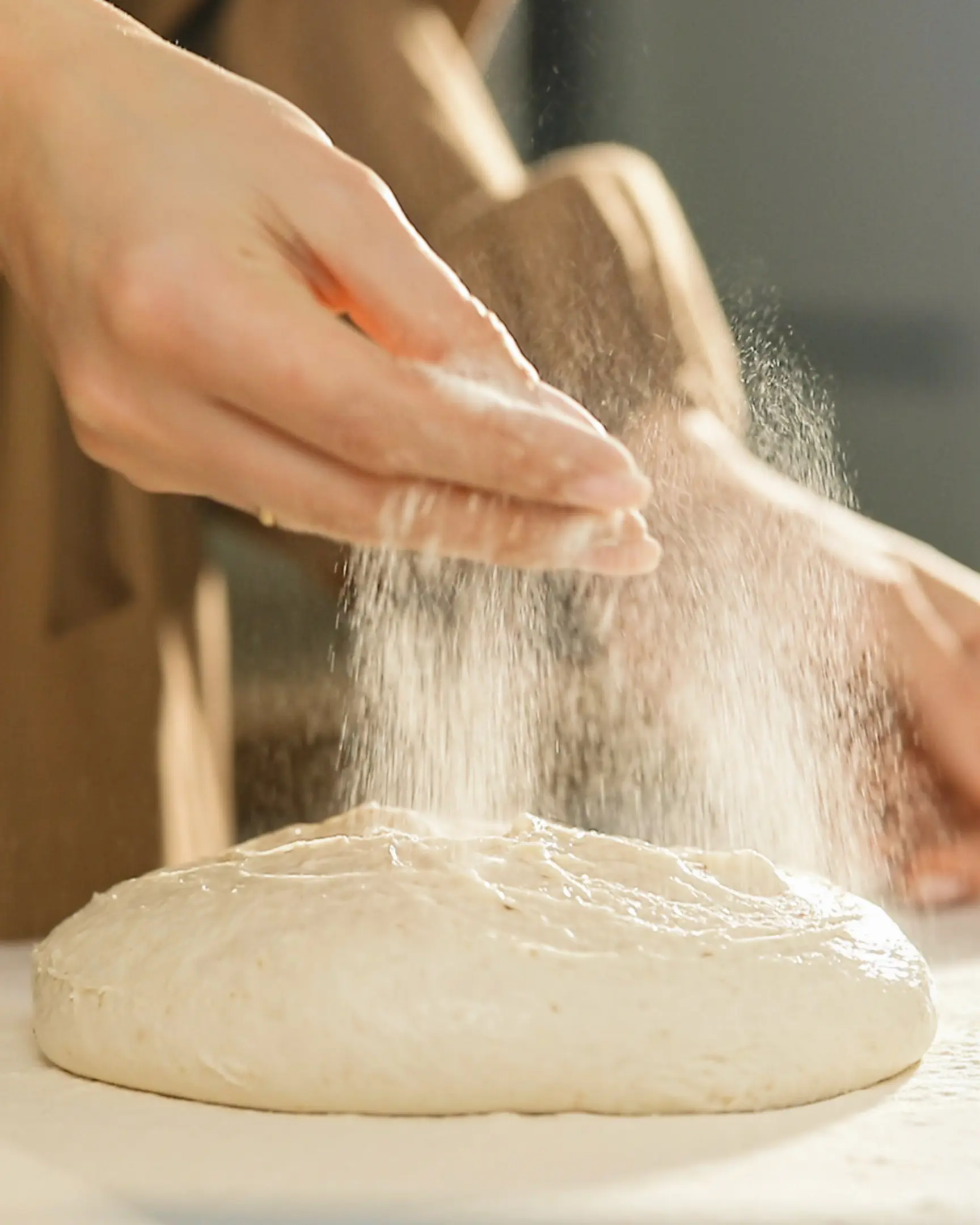 Hands dusting wheat flour onto elastic baguette dough for shaping, an essential step in the preparation recipe.